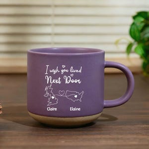 May include: A purple ceramic mug with a tan base. The mug features the text "I wish you lived Next Door" with a map of the UK and the USA. The names "Claire" and "Elaine" are below the maps.