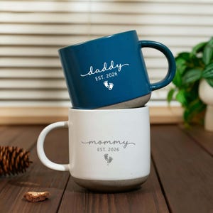 May include: A dark blue ceramic mug stacked on a white ceramic mug. The blue mug features the white text "daddy EST. 2026" with a baby footprint icon. The white mug features the gray text "mommy EST. 2026" with a baby footprint icon below the text.