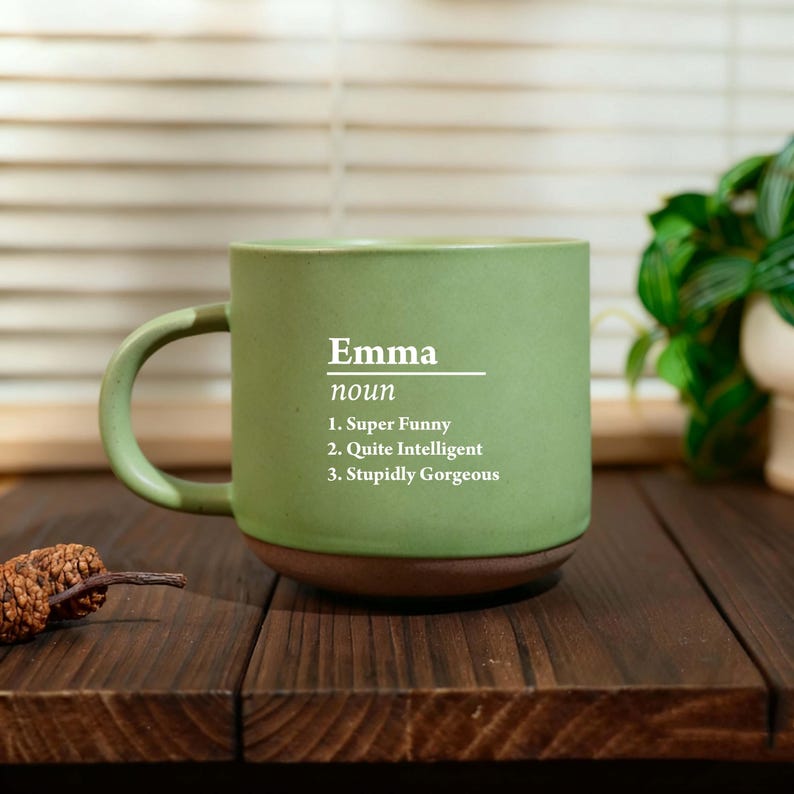 May include: A green ceramic mug with a brown base and handle. The mug features the name "Emma" with the definition of a noun, followed by three humorous descriptions: "Super Funny," "Quite Intelligent," and "Stupidly Gorgeous."