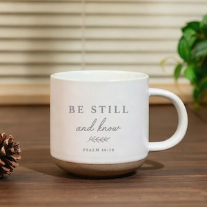 Op de afbeelding: Een witte keramische mok met een handvat, voorzien van de grijze tekst "BE STILL and know", een kleine bladillustratie en de vermelding "PSALM 46:10". De onderkant van de mok heeft een natuurlijke, ongeglazuurde, gespikkelde afwerking.