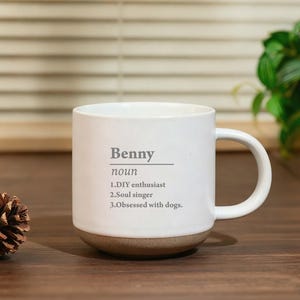 May include: White ceramic mug with a brown base and a curved handle. The mug features the name "Benny" and the word "noun" printed in gray, along with three descriptive phrases: "DIY enthusiast," "Soul singer," and "Obsessed with dogs."