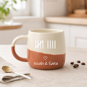 Peut inclure: Un mug en céramique bicolore avec une partie supérieure crème et une partie inférieure terracotta. Le mug présente un graphique de marques de comptage blanc, une petite icône de cœur et le texte imprimé "Noah & Sara" sur la face avant.