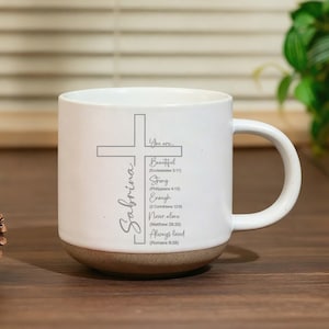 Peut inclure: Mug en céramique blanche avec une base de couleur naturelle. Le mug présente une croix et le nom "Sabrina" en écriture cursive. Le texte comprend "You are... Beautiful, Strong, Enough, Never alone, Always loved" avec des références bibliques.