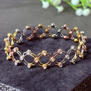 Puede incluir: Una pulsera de metal tricolor con un patrón repetitivo de eslabones circulares. Los eslabones alternan tonos dorados, plateados y oro rosa, con pequeñas piedras transparentes incrustadas en las uniones de cada segmento circular del diseño.