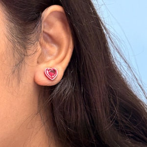Può includere: Primo piano di un orecchino a forma di cuore. L'orecchino presenta una gemma rosa vibrante incastonata in un metallo color argento. L'orecchino è indossato su un orecchio, con capelli castano scuro visibili.