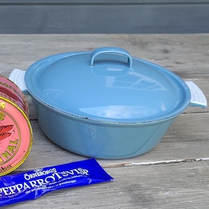 Könnte beinhalten: Eine hellblaue ovale Auflaufform mit Deckel, eine rosa Dose "ABBA ANSJOVIS" und eine blaue Tube "Pepparrot Svisp" auf einer Holzoberfläche. Die Dose hat eine rote Abbildung eines Flusskrebses.