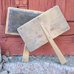 Könnte beinhalten: Zwei antike Wollkarden aus Holz, eine mit dunkelgrauer Oberfläche, lehnen an einer verwitterten roten Holzwand. Die Karden sind auf einer Steinoberfläche platziert, was ein rustikales, Vintage-Thema suggeriert. Die Holzwerkzeuge weisen Gebrauchsspuren auf.