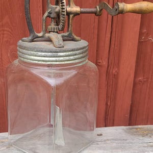 May include: A vintage glass butter churn featuring a square-shaped clear glass jar with a metal lid. The top mechanism is made of dark, weathered cast iron with a gear system and a wooden handle attached to a crank for manual operation.