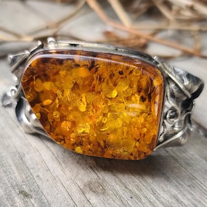 Könnte beinhalten: Ein silbernes Armband mit einem großen, rechteckigen Bernstein. Der Bernstein hat eine warme, goldgelbe Farbe mit sichtbaren Einschlüssen. Die Silberfassung hat ein verziertes, florales Design. Das Armband wird auf einer verwitterten Holzoberfläche präsentiert.