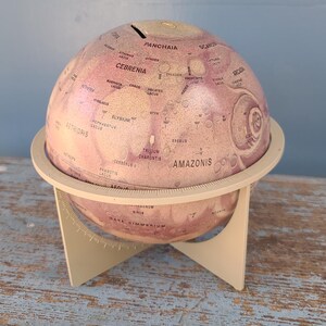 Könnte beinhalten: Eine Geldspardose in Form eines Vintage-Globus mit rosa-goldener Oberfläche. Der Globus zeigt geografische Details und ist auf einem cremefarbenen Ständer montiert. Er hat einen Schlitz zum Einwerfen von Münzen.