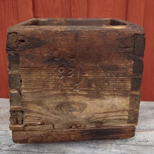 Può includere: Una scatola quadrata in legno usurato con venature visibili e toni marrone scuro. Il pannello frontale presenta i numeri 1231 e 2 stampati. La costruzione mostra giunti a pettine esposti agli angoli, indicando un design rustico e utilitario.