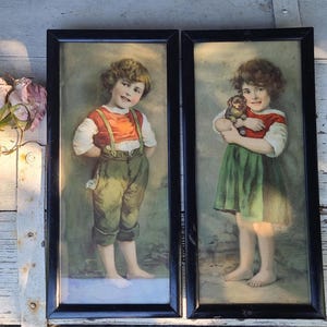 Könnte beinhalten: Zwei gerahmte Vintage-Drucke. Ein Druck zeigt einen jungen Mann in einem orangefarbenen Hemd, grünen Latzhosen und Hosenträgern. Der andere zeigt ein junges Mädchen in einem rot-weißem Hemd und grünem Kleid, das ein kleines Stofftier hält. Beide Drucke haben schwarze Rahmen.