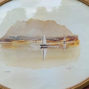 Könnte beinhalten: Ein ovales Wandbild mit einem Aquarellgemälde eines Segelboots auf ruhigem Wasser vor einer Bergkulisse. Das Kunstwerk ist in einem dünnen, strukturierten, goldfarbenen Metallrahmen mit einer perlenartigen Kantenverzierung eingefasst.