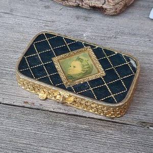 Può includere: Un portagioie rettangolare antico con un motivo trapuntato nero e finiture dorate, con una piccola immagine incorniciata. La scatola è circondata da conchiglie, un riccio di mare e un orologio in legno su una superficie di legno invecchiato.
