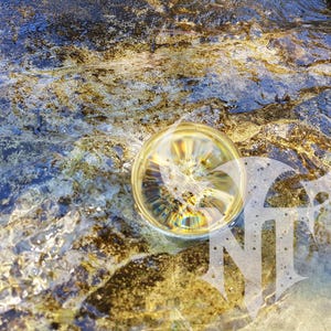 Könnte beinhalten: Eine klare, kugelförmige Glaskugel liegt untergetaucht in flachem, fließendem Wasser über einer texturierten, goldbraunen Felsoberfläche. Ein weißes, stilisiertes Logo mit den Buchstaben NT ist unten rechts im Bild eingeblendet.