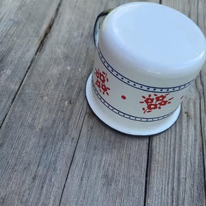 May include: White enamel mug with a blue rim and handle, decorated with red floral patterns and a blue dotted border. The mug sits on a weathered wooden surface.