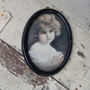 Puede incluir: Retrato ovalado enmarcado en negro de un niño con cabello rubio rizado. El niño tiene una expresión serena, con las manos juntas. El retrato está sobre una superficie de madera desgastada pintada de blanco.
