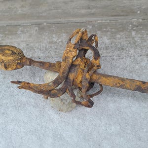 Könnte beinhalten: Ein antikes, rostiges Metallschwert mit detailliertem Knauf und langer, schlanker Klinge. Das Schwert hat eine verwitterte braune und orange Farbe, was auf Alter und Witterungseinflüsse hindeutet. Es liegt auf einer hellgrauen Oberfläche.