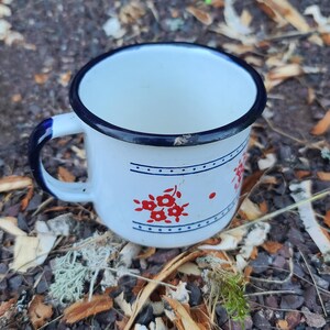 May include: White enamel mug with a dark blue rim and handle. The mug features a red floral design and a dark blue stripe. The mug is sitting on a bed of mulch and small rocks.