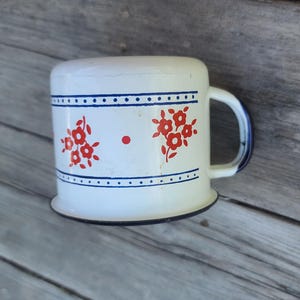 May include: White enamel mug with red floral designs and blue trim. The mug has a rounded top and a handle with blue accents. The mug is sitting on a weathered wooden surface.