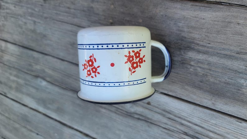 May include: White enamel mug with red floral designs and blue trim. The mug has a rounded top and a handle with blue accents. The mug is sitting on a weathered wooden surface.