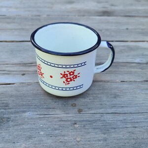May include: White enamel mug with a blue rim and handle, decorated with red floral patterns and blue dotted lines. The mug sits on a weathered wooden surface, showcasing a vintage aesthetic.