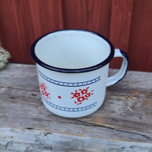 May include: White enamel mug with a blue rim and handle. The mug features a red floral design and a blue dotted border. The mug is sitting on a weathered wooden surface.