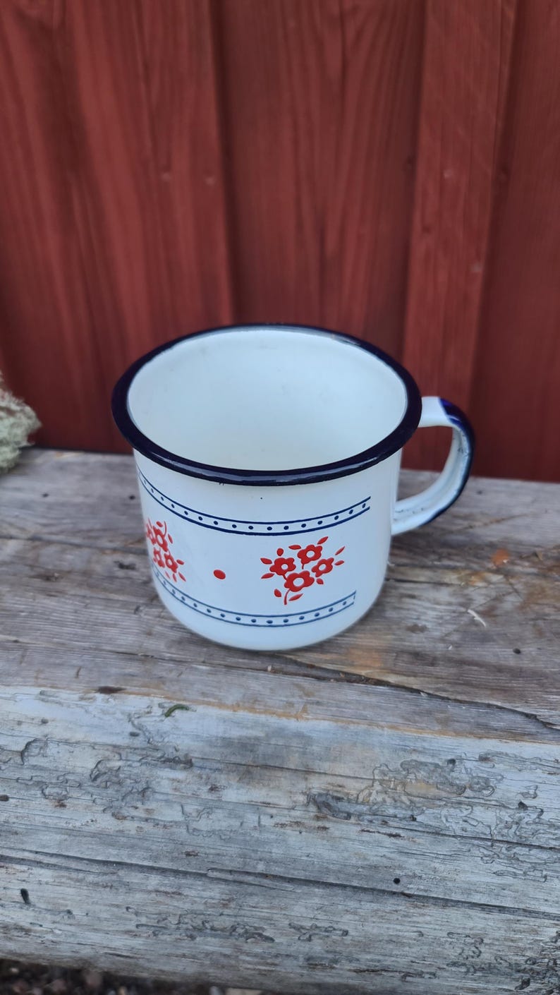 May include: White enamel mug with a blue rim and handle. The mug features a red floral design and a blue dotted border. The mug is sitting on a weathered wooden surface.