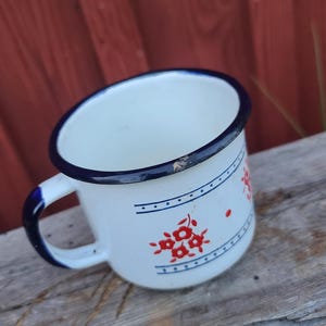 May include: White enamel mug with a blue rim and handle. The mug features a red floral design and blue horizontal lines with dots. The mug is sitting on a weathered wooden surface.
