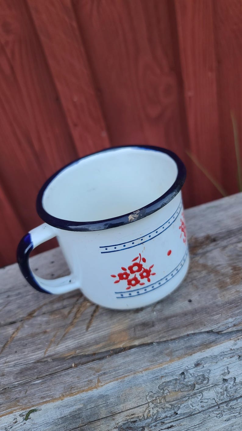 May include: White enamel mug with a blue rim and handle. The mug features a red floral design and blue horizontal lines with dots. The mug is sitting on a weathered wooden surface.
