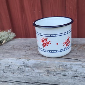 May include: White enamel mug with a blue rim and a floral design in red. The mug has a blue band with red floral accents and is set on a weathered wooden surface. A classic camping or kitchenware item.