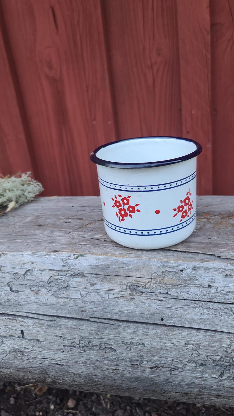 May include: White enamel mug with a blue rim and a floral design in red. The mug has a blue band with red floral accents and is set on a weathered wooden surface. A classic camping or kitchenware item.