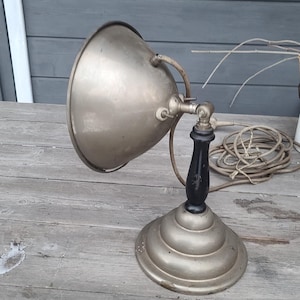 May include: An antique silver-toned desk lamp with a black handle. The lamp has a rounded shade and a circular base. The lamp is sitting on a weathered wooden surface. A coiled electrical cord is visible in the background.