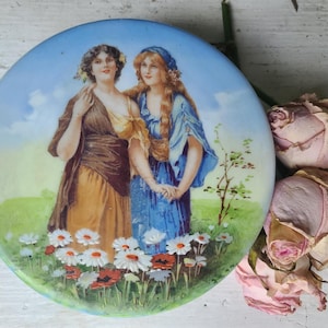 Könnte beinhalten: Runder, antiker Porzellanteller mit einer gemalten Szene von zwei Frauen in langen Kleidern in einem Blumenfeld. Der Teller hat einen blauen Himmel als Hintergrund. Getrocknete rosa Rosen sind um den Teller angeordnet.