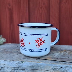 May include: White enamel mug with a blue rim and handle. The mug features red floral designs and blue dotted lines. The mug is sitting on a weathered wooden surface with a red background.