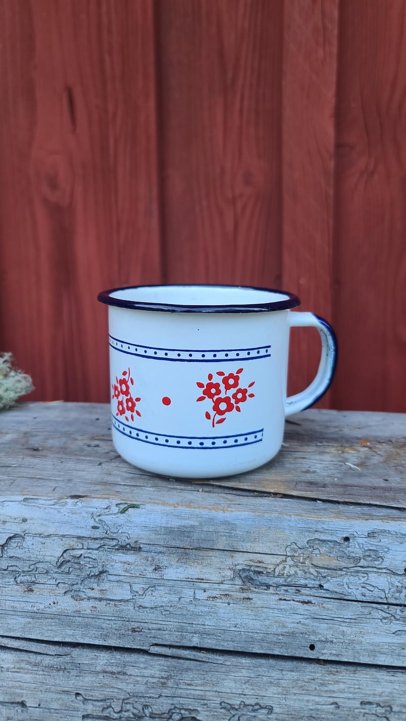 May include: White enamel mug with a blue rim and handle. The mug features red floral designs and blue dotted lines. The mug is sitting on a weathered wooden surface with a red background.