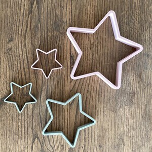 May include: Four star-shaped cookie cutters in varying sizes. The cutters are in pastel colors: light pink and light green. The cutters are made of plastic and are arranged on a wooden surface.