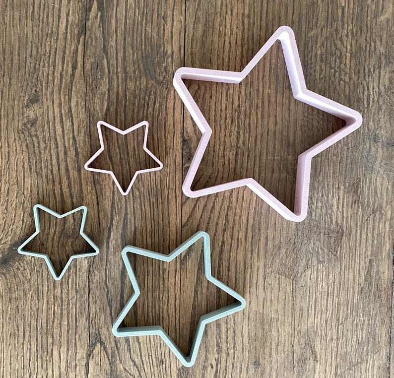 May include: Four star-shaped cookie cutters in varying sizes. The cutters are in pastel colors: light pink and light green. The cutters are made of plastic and are arranged on a wooden surface.