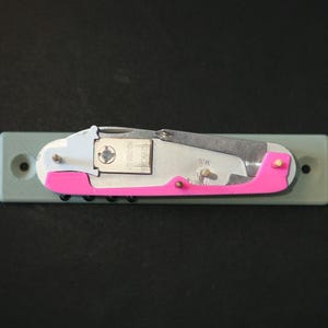 Puede incluir: Una multiherramienta con una hoja plateada y un mango rosa brillante montada sobre una base verde claro. La herramienta tiene un pequeño componente plateado con el texto "128GB" y otras marcas.