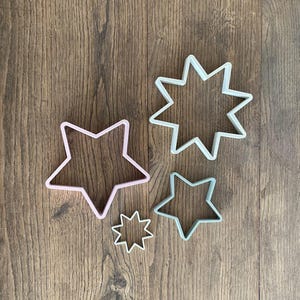 May include: Four star-shaped cookie cutters in varying sizes. The cutters are in pastel colors: pink, light green, and white. The cutters are made of plastic and are arranged on a wooden surface.