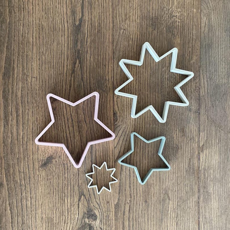 May include: Four star-shaped cookie cutters in varying sizes. The cutters are in pastel colors: pink, light green, and white. The cutters are made of plastic and are arranged on a wooden surface.