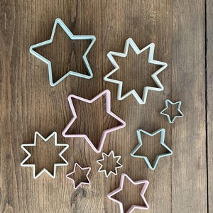 May include: A collection of star-shaped cookie cutters in various sizes and designs. The cutters are in shades of pink and mint green, with both classic five-point and multi-point star shapes. They are arranged on a wooden surface.