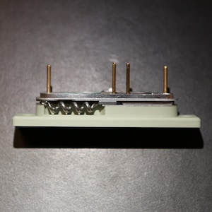 May include: A small, rectangular, pale green electronic component with four gold-colored pins. A silver metal plate is attached, with a coiled spring visible. The component is set against a dark gray background.