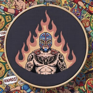 May include: A cross-stitch embroidery of a masked wrestler in a blue and red mask, surrounded by flames. The wrestler has tattoos and is set against a dark background within a wooden hoop. The background is a pattern of wrestling masks and figures.