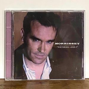 Puede incluir: Una caja de CD con Morrissey y el álbum "Vauxhall and I". La portada muestra un primer plano del rostro de Morrissey, con el nombre del artista en blanco. La caja es transparente con un lomo morado.