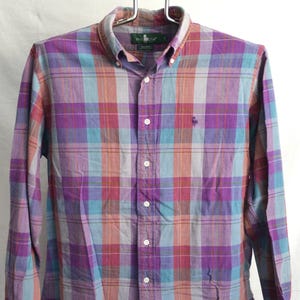 May include: A long-sleeve button-down shirt featuring a plaid pattern in shades of purple, blue, and orange. The shirt has a button-down collar, a front button placket, and a small embroidered logo on the left chest area.