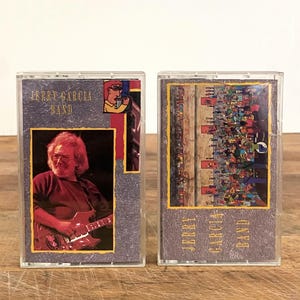 Könnte beinhalten: Zwei Vintage-Kassettenbänder mit der Jerry Garcia Band. Auf einem Band ist ein Foto von Jerry Garcia beim Gitarrespielen zu sehen, auf dem anderen eine farbenfrohe Illustration einer Menschenmenge. Beide Bänder tragen den Namen der Band.