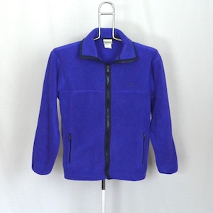 Peut inclure: Une veste en polaire violette avec une fermeture éclair et deux poches zippées. La veste a un col noir et un petit logo sur la poitrine gauche.
