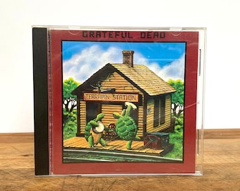 May include: A red CD case with a picture of a small wooden building with the words "TERRAPIN STATION" on the front. Two green turtles are playing music in front of the building. The text "GRATEFUL DEAD" is at the top of the case.