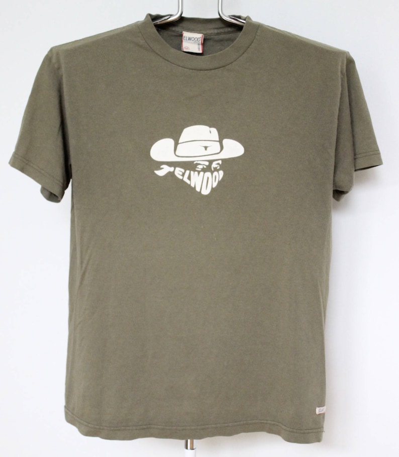 Op de afbeelding: Een groen T-shirt met korte mouwen met een witte afbeelding van een cowboy met een hoed en het woord "ELWOOD" gedrukt op de voorkant.